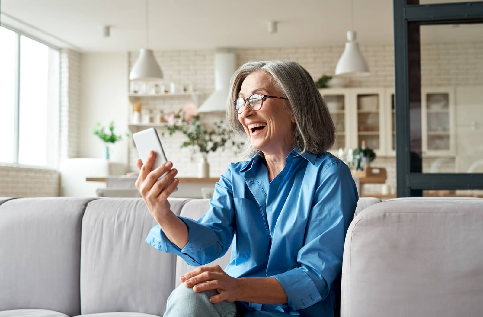 An older adult on the couch in assisted living laughing while holding their cell phone up to their face.