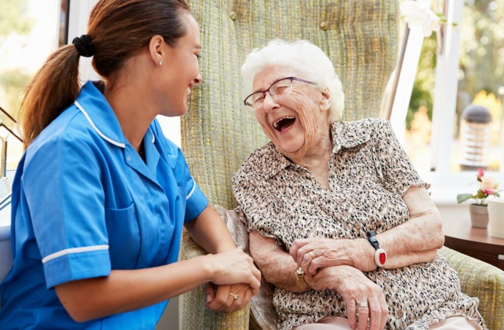 A senior woman sitting in a chair and laughing while having a conversation with a staff member from her senior living community.