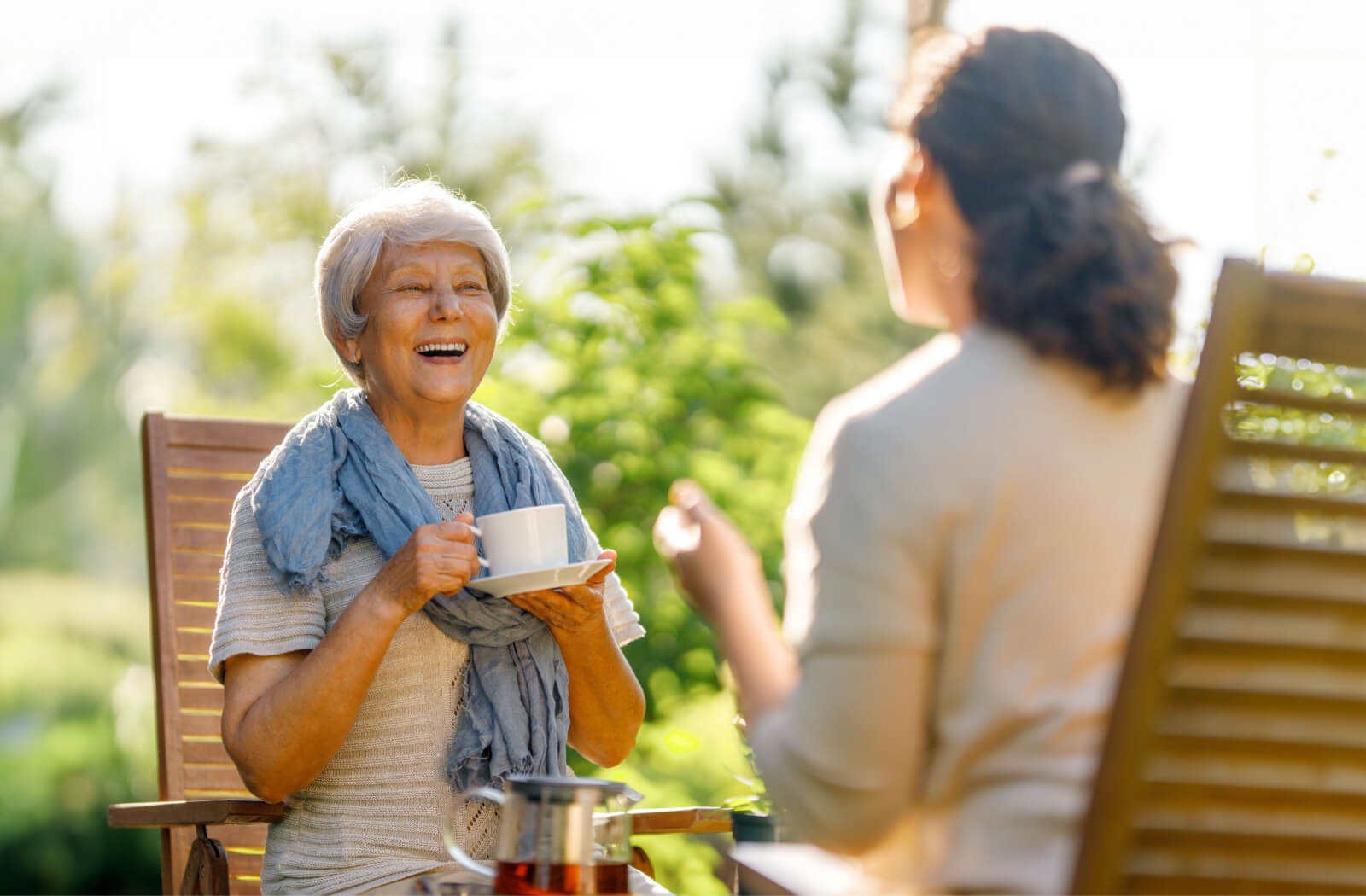 A senior woman and her daughter sitting on lawn chairs outside, smiling and talking to each other while holding a cup of tea