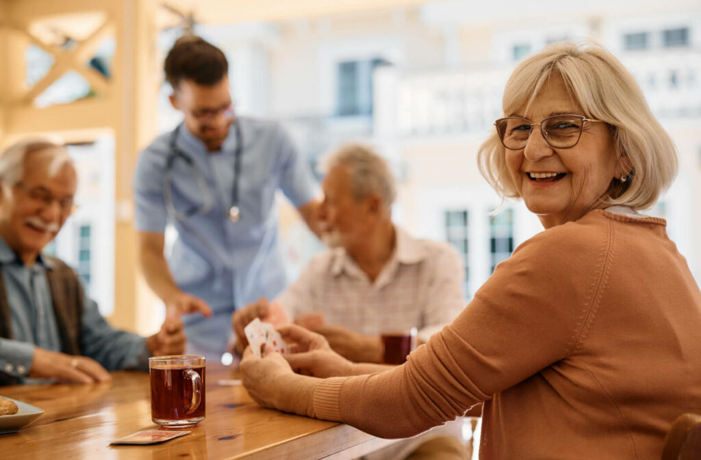 A group of seniors at a senior living community sit around a table laughing and playing cards together