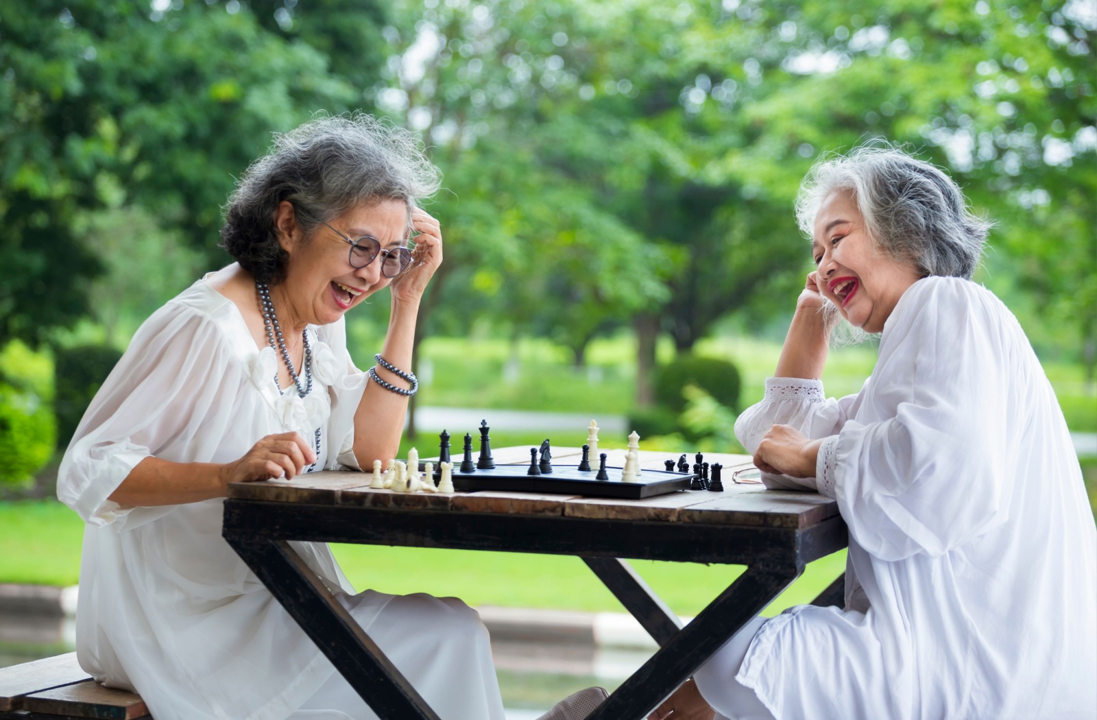 A couple of seniors laugh together as they play a game of chess in an outdoor sitting area.