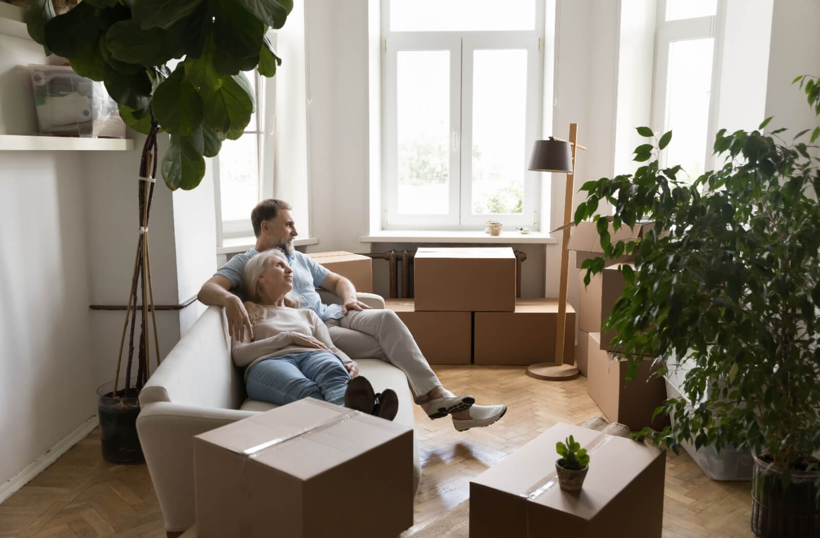 An older couple resting together on a couch surrounded by moving boxes in their packed-up home.