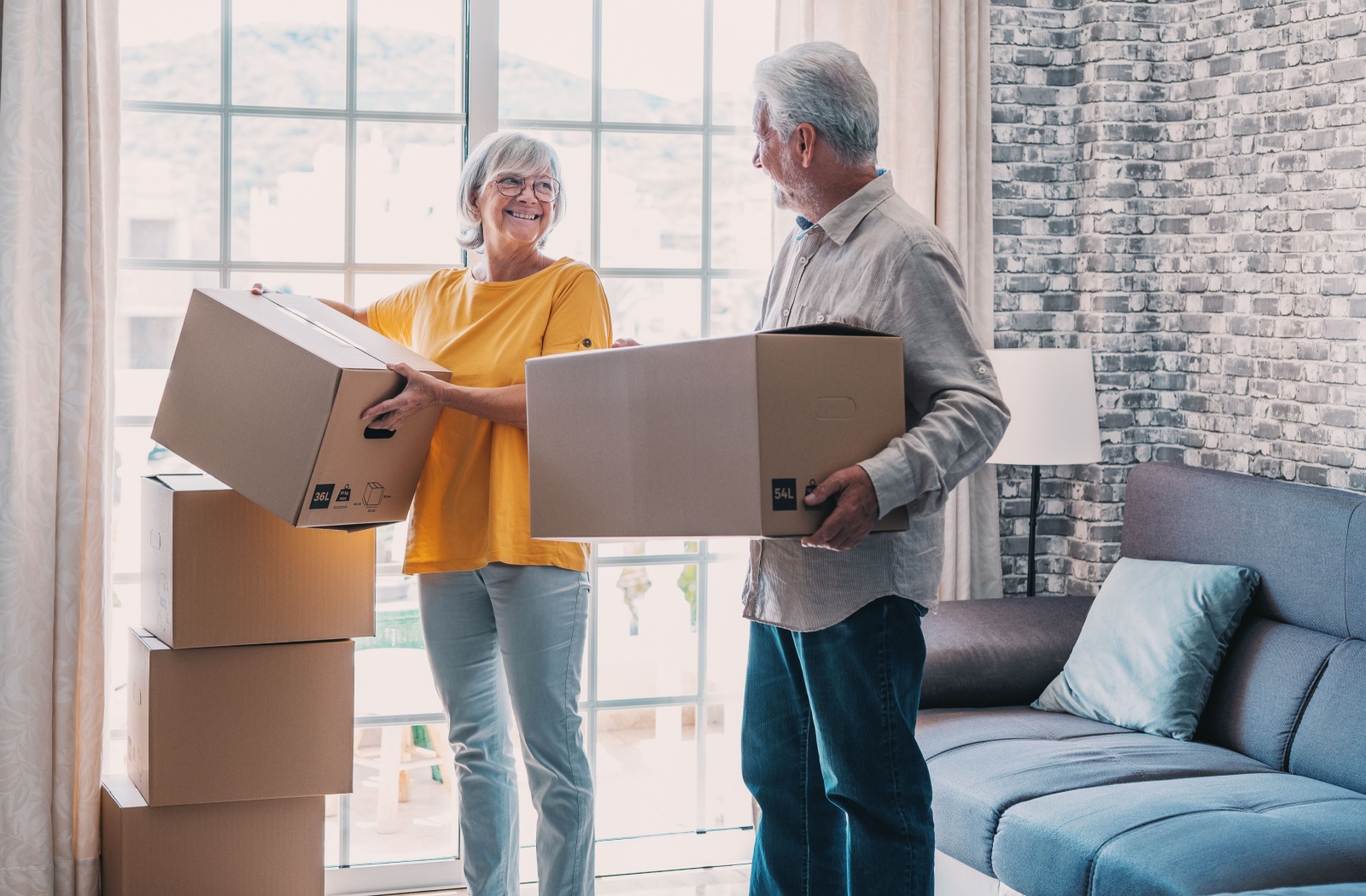 An older couple smiling at one another while stacking moving boxes in their sunlit living room.