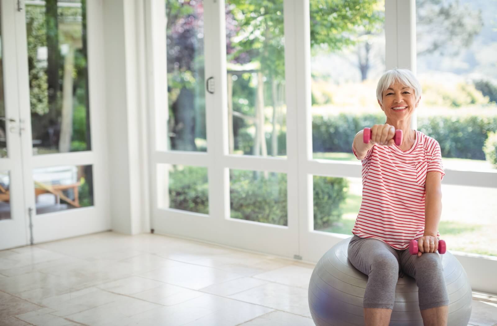 A senior resident sits on an exercise ball, holding light weights for arm exercise.
