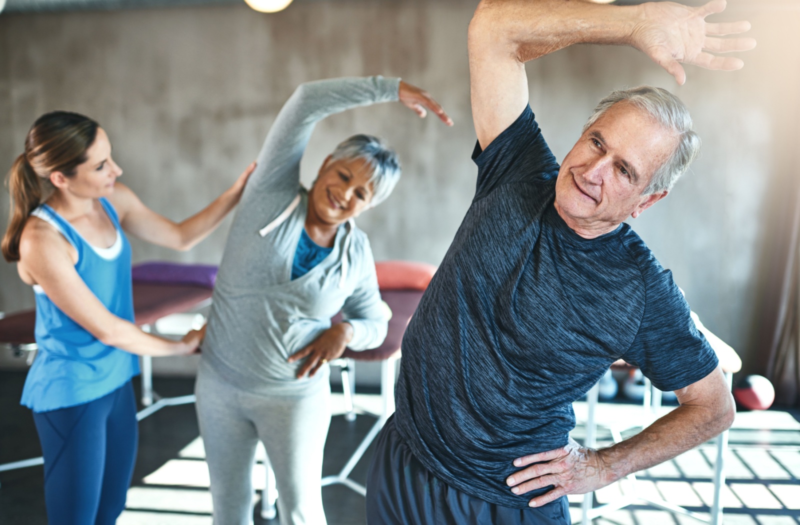 Two older adults, assisted by a fitness instructor, raise their right arms above their heads to stretch the right side of their torsos.