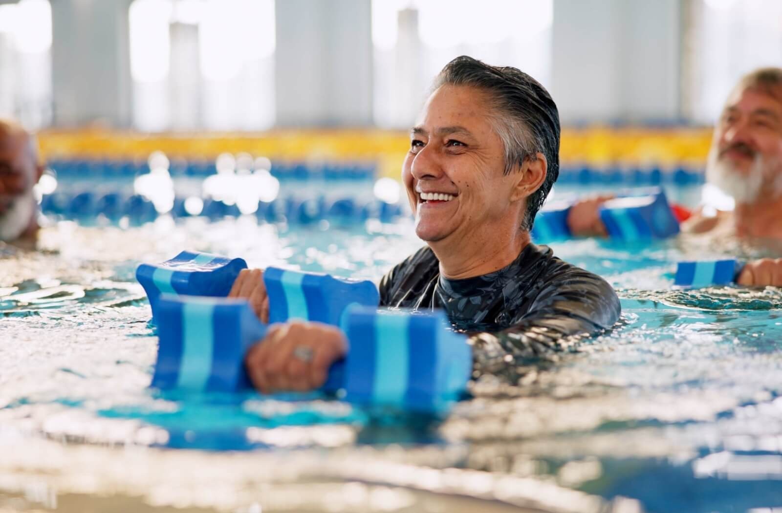 A smiling older adult works out in a swimming pool during an aquatic therapy class at a senior living community.